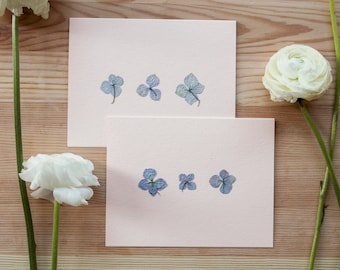 Peut inclure: Deux cartes roses avec des fleurs bleues séchées disposées en rangées. Les cartes sont sur une surface en bois clair.