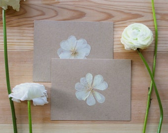 Peut inclure: Deux cartes brunes avec des fleurs blanches pressées sur un fond de bois clair. Les fleurs sont délicates et ont un aspect naturel et rustique.