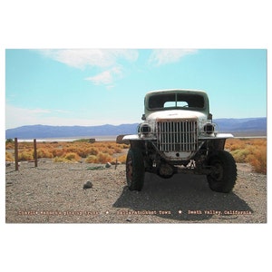 May include: A white, weathered pickup truck sits in a desert landscape. The truck has a rusty grill and a large, flat bed. The truck is parked in front of a sign that reads "Ballarat Ghost Town" and "Death Valley, California".
