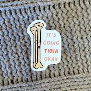 May include: A white sticker with a cartoon drawing of two broken bones hugging each other. The text on the sticker reads "It's going tibia okay."