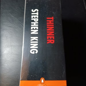 May include: A black and orange book cover with the title "Thinner" and the author "Stephen King" in white text. The Penguin Audiobooks logo is at the bottom of the cover.