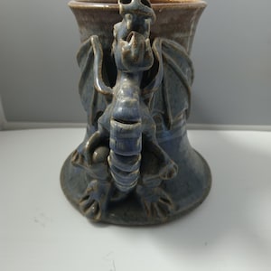May include: A blue ceramic mug with a dragon design. The mug features a detailed dragon sculpture wrapped around the body, with wings and a head at the top. The rim is a darker brown color.