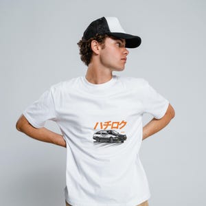Puede incluir: Camiseta blanca con una ilustración de un coche blanco y negro y el texto japonés "ハチロク" impreso en la parte delantera. La persona lleva una gorra de camionero blanca y negra.