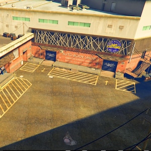 Può includere: Vista aerea di un'officina di carrozzeria con un'ampia area di parcheggio. L'edificio ha un'insegna con la scritta "CUSTOMS" e "AUTO BODY SHOP". Linee gialle segnano i parcheggi. La scena è immersa nella luce del sole.