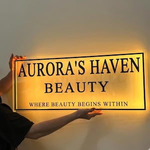May include: Illuminated sign with the text "AURORA'S HAVEN BEAUTY" in black, with the tagline "WHERE BEAUTY BEGINS WITHIN." The sign has a yellow glow and is held by a person.