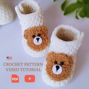 Peut inclure: Une paire de chaussons en crochet blancs avec un motif de visage d'ours brun sur le devant. Les chaussons ont un aspect doux et texturé. Le texte "CROCHET PATTERN VIDEO TUTORIAL" est également présent.
