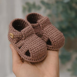 May include: A pair of handmade, crochet baby sandals in a muted mauve color. The sandals feature an open-toe design with a strap across the top and a small wooden button closure. The texture of the crochet stitch is clearly visible.