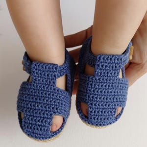 May include: A pair of blue crochet baby sandals. The sandals have a woven design with a strap and button closure. The soles are a light tan color. The sandals are being held by a person.