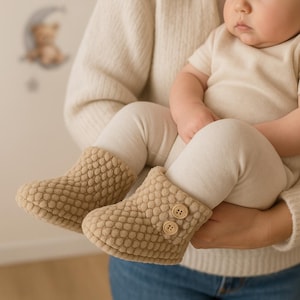 Op de afbeelding: Een paar beige babyschoentjes met een textuur, bubbelachtig patroon. Elke schoen heeft twee kleine, ronde knopen. De schoentjes zijn ontworpen om de voetjes van een baby warm en comfortabel te houden.