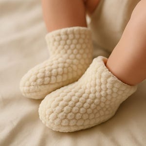 Op de afbeelding: Close-up van twee cr&egrave;mekleurige babyslofjes met een textuur, bubbelachtig patroon. De slofjes liggen op een zacht, neutraal getint oppervlak, wat duidt op comfort en warmte. De afbeelding benadrukt het gezellige ontwerp.