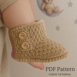Op de afbeelding: Een beige babybootie met een getextureerd oppervlak en twee houten knopen. De bootie is gemaakt van een zacht, pluche materiaal. De tekst "PDF Pattern VIDEO TUTORIAL" staat onderaan de afbeelding.