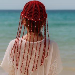 May include: A red crocheted head covering with beaded fringe. The head covering is a deep red color and has a net-like design. Long strands of red yarn are adorned with white beads, creating a flowing, decorative effect.