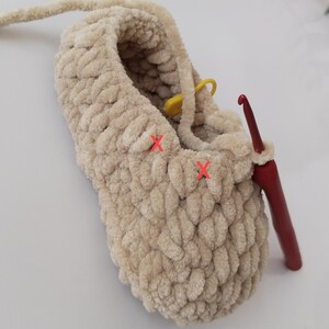 Op de afbeelding: Een beige, zachte pantoffel in wording, met een rode haaknaald erin. Een gele steekmarkeerder is aan het garen bevestigd. De pantoffel is gemaakt van dik, getextureerd garen en de steken zijn zichtbaar.