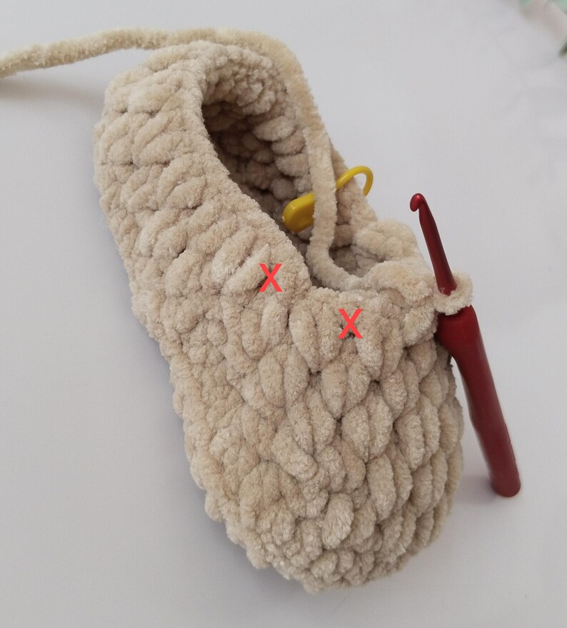 Op de afbeelding: Een beige, zachte pantoffel in wording, met een rode haaknaald erin. Een gele steekmarkeerder is aan het garen bevestigd. De pantoffel is gemaakt van dik, getextureerd garen en de steken zijn zichtbaar.