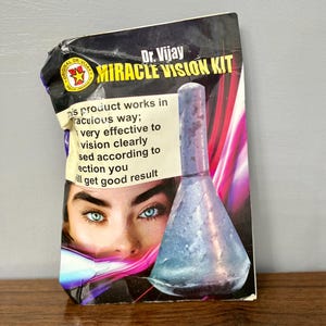 Może przedstawiać: Fioletowo-różowe opakowanie z srebrną i niebieską szklaną butelką. Opakowanie zawiera tekst "Dr. Vijay Miracle Vision Kit" i "Ten produkt działa w cudowny sposób; bardzo skuteczny, aby widzieć wyraźnie, stosowany zgodnie z instrukcjami, uzyskasz dobre rezultaty".