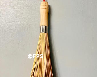 May include: A wooden handled bamboo whisk with a red string hanging from the top. The whisk is hanging on a gray wall.
