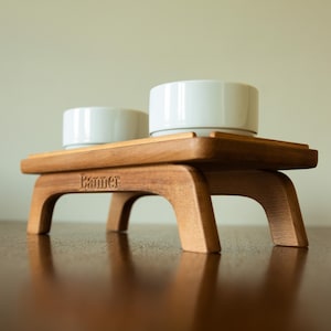 May include: A wooden pet bowl stand with two white ceramic bowls. The stand is made of light brown wood and has the word "Banner" engraved on the front. The bowls sit on a raised platform.
