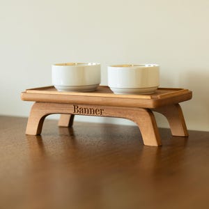 May include: A wooden serving tray with two white bowls with gold rims. The tray has a rectangular top and curved legs. The word "Banner" is engraved on the front of the tray.
