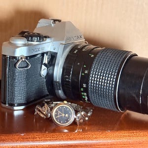May include: A vintage Pentax ME Super 35mm film camera with a black and silver body and a long black lens. A small, gold-toned watch with a black face and gold hands rests in front of the camera. The watch has a decorative band.