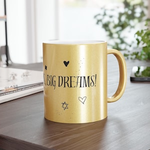 Op de afbeelding: Gouden koffiemok met zwarte tekst "Go Big Dreams!" met een hart- en stervormig ontwerp.