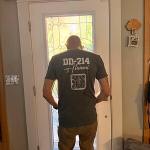 May include: A dark gray short-sleeved t-shirt featuring white text on the back that reads DD-214 Alumni, positioned above a square black and white QR code. The shirt is worn with tan trousers, and the design is centered on the upper back area.