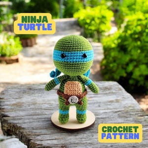 May include: Crochet pattern for a green, blue, and brown Ninja Turtle amigurumi doll. The doll is standing on a wooden base and has the text "NINJA TURTLE" and "CROCHET PATTERN" on the image.