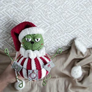 Op de afbeelding: Een groen gebreid Grinch-achtig personage dat een rood-wit gestreepte hoed en een rood-wit gestreepte jurk draagt. Het personage houdt een touwtje met kleine vlaggetjes met kerstthema's vast.
