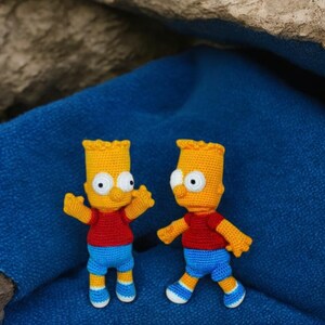 Puede incluir: Dos figuras de ganchillo hechas a mano de un personaje de dibujos animados. Las figuras tienen piel amarilla, camisas rojas, pantalones cortos azules y zapatos blancos y azules. Están posadas sobre un fondo de tela azul.