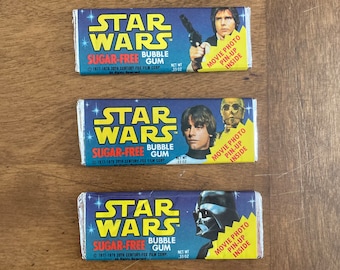 May include: Three vintage Star Wars bubble gum wrappers with a blue background and white text. Each wrapper features a different character from the film: Han Solo, Luke Skywalker, and Darth Vader. The text on the wrappers reads "Star Wars Sugar-Free Bubble Gum" and "Movie Photo Pin-Up Inside".