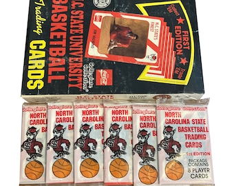 May include: A box of 1st edition North Carolina State University Basketball trading cards. The box is black with red and white text. The cards are in individual plastic packs with a red and white mascot logo.