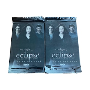 以下が含まれることがあります： 「トワイライト・サーガ：エクリプス」のプレミアムトレーディングカードの2つの密封パック。各パックは、キャラクターの画像と「eclipse」のタイトルが付いた暗い背景が特徴です。パックには「1パックあたり6枚のカード」とNECAのロゴが記載されています。
