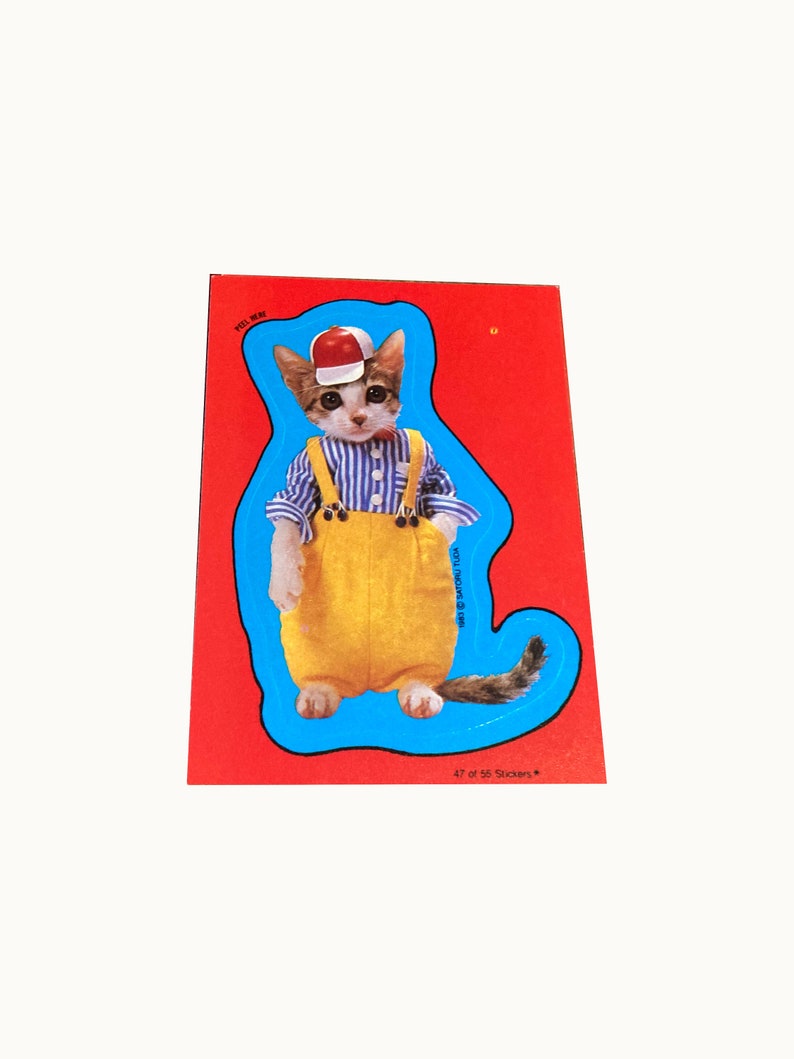 May include: A sticker featuring a cartoon cat wearing a blue and white striped shirt, yellow overalls, and a red baseball cap. The cat is standing on a blue background with a white outline. The sticker is part of a set of 56 stickers, and this is number 47.