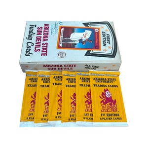 Op de afbeelding: Een witte doos met Arizona State Sun Devils trading cards met de woorden "First Edition" en "All-Time Greats". Verschillende ongeopende pakjes kaarten liggen voor de doos, elk met het ASU-logo en de woorden "1st Edition 8 Player Cards."