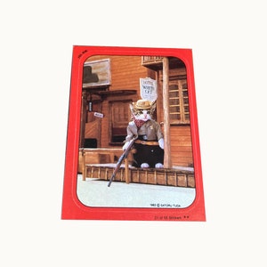 May include: A white cat wearing a cowboy hat and a sheriff's badge stands in front of a wooden building with a sign that reads "Hotel White Cat". The cat is holding a rifle and is looking to the right. The image is from a sticker collection and is labeled "21 of 55 Stickers".