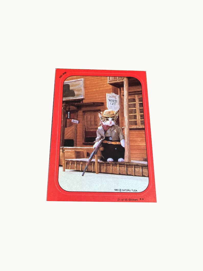 May include: A white cat wearing a cowboy hat and a sheriff's badge stands in front of a wooden building with a sign that reads "Hotel White Cat". The cat is holding a rifle and is looking to the right. The image is from a sticker collection and is labeled "21 of 55 Stickers".