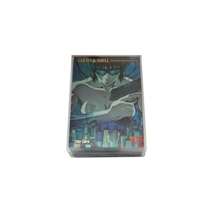 Peut inclure: Une carte de titre rectangulaire Ghost in the Shell représentant un personnage aux cheveux sombres et aux lunettes teintées de bleu tenant une arme de poing. L'arrière-plan montre un paysage urbain bleu. Le texte indique Ghost in the Shell.