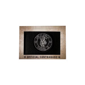 May include: Black leather patch with a white embroidered design featuring a skull with a scythe, the words "SONS OF ANARCHY" and "CHARMING, CA". The patch is set against a brown background with the text "OFFICIAL CONTRABAND".