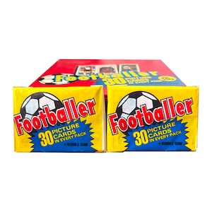 May include: Two yellow rectangular packs of Footballer bubble gum and picture cards, each featuring a black and white soccer ball graphic, the red text Footballer, and blue text stating 30 picture cards in every pack plus bubble gum.