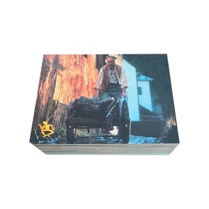 May include: An older man in a brown vest and a cap pushes a wooden wheelbarrow down a cobblestone street. The wheelbarrow is filled with logs. The scene is lit by the warm glow of the setting sun.