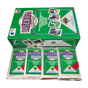 May include: A green baseball card box set with the text "BASEBALL 1990 EDITION" and several unopened packs. The box features baseball cards and the MLB logo. The packs are silver and green.