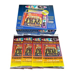 Op de afbeelding: Een doos met "Silly CDs" verzamelkaarten met meerdere pakjes. De doos en de pakjes tonen de parodie van de albumhoes "NStink". Elk pakje bevat 5 kaarten en willekeurig ingevoegde stickers. Op de doos staat "Premier Edition".