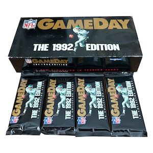 Peut inclure: Un coffret noir de cartes à collectionner NFL Gameday 1992. La boîte a des lettres dorées et une image d'un joueur de football américain. Quatre paquets de cartes non ouverts sont devant la boîte. Chaque paquet porte le logo Gameday et le texte "The 1992 Edition".