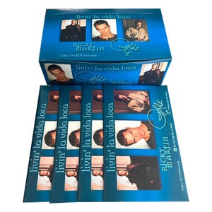 May include: A blue box with a photo of Ricky Martin on the front. The box says "livin' la vida loca" and "Ricky Martin". There are four blue packs of photocards stacked on top of the box. Each pack has a photo of Ricky Martin and the text "livin' la vida loca".