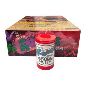 May include: A cylindrical red and white container labeled "Players Kit" with the text "Since the... Beginning of Time" and "Roks" in front of a rectangular game box featuring a prehistoric landscape with a pterodactyl and a volcano illustration.