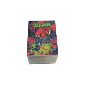 May include: A stack of rectangular trading cards featuring the Spawn comic book character. The top card displays the green Spawn logo, a dark figure in a flowing red cape, and swirling green energy effects against a dark, dramatic background.