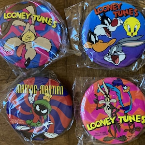 Könnte beinhalten: Vier runde, bunte Plastikknöpfe mit Figuren aus der Zeichentrickserie Looney Tunes. Die Knöpfe sind rosa, blau, rot und schwarz mit weißen Buchstaben. Die Figuren sind Wile E. Coyote, Road Runner, Marvin der Marsmensch und Bugs Bunny.