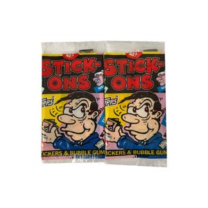 May include: Two sealed packs of Topps Stick-Ons stickers and bubble gum. The packaging features a cartoon man holding a small card, with the words "STICK-ONS" in red and black. The bottom of the pack reads "STICKERS & BUBBLE GUM".