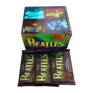 Könnte beinhalten: Eine Display-Box und drei versiegelte Folienpackungen mit The Beatles Sammelkarten. Die Box zeigt ein buntes, psychedelisches Raster mit Porträts der Bandmitglieder und den Schriftzug THE BEATLES. Jede schwarze Folienpackung zeigt ebenfalls THE BEATLES.