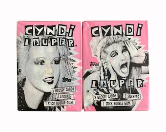 May include: Two pink and black trading card packs featuring Cyndi Lauper. Each pack includes 3 glossy cards, 3 stickers, and 1 stick of bubble gum.