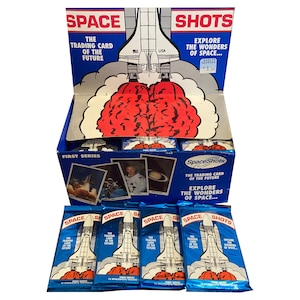 May include: A display box of "Space Shots" trading cards, featuring a blue and white design with a rocket ship graphic. The box is open, revealing individual card packs and sample cards with space-themed images. The text reads "Explore the Wonders of Space."